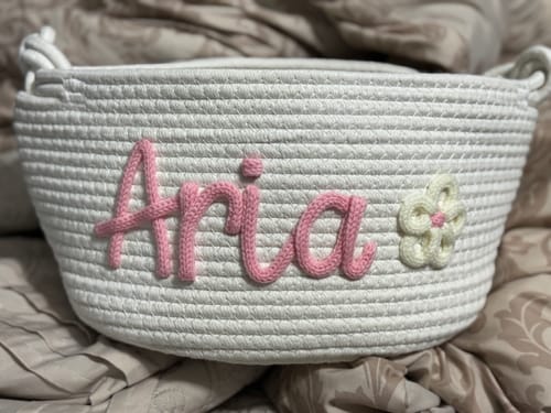 Customer's personalized knitted basket in white, featuring the name 'Aria' in pink yarn and a knitted flower icon.