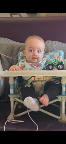 A baby sits in the reviewer's light blue checkered Harper baby chair v2 placed on a purple couch with its tray attached.