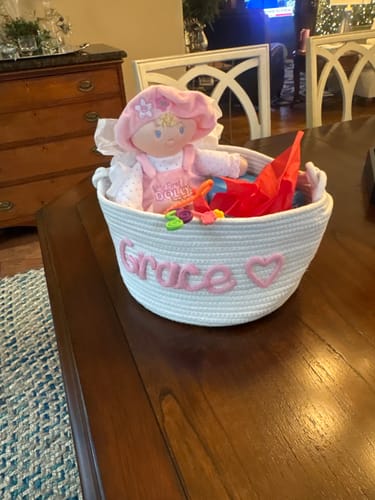 Customer's white personalized knitted basket with the name "Grace" and a heart icon, filled with toys and a doll.