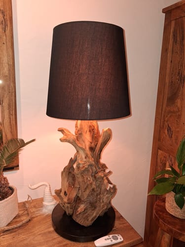 Ein Kunde zeigt die eingeschaltete Wurzelholz-Tischlampe mit rustikalem Holzfuß und schwarzem Schirm auf einem Tisch.