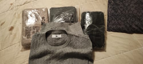Customer's grey cable-knit pullover laid out on a bed, with two other sweaters still in their plastic packaging.