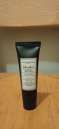 Recensent toont de zwarte tube Istanbul Handcrème, staand op een houten ondergrond.
