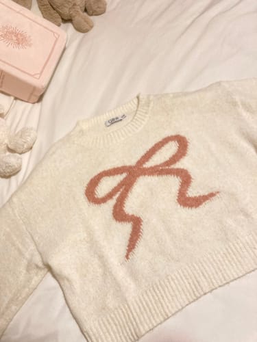 Reviewer's cream knit jumper with a large, pink embossed bow shown laid flat on a bed.