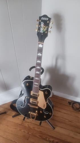 Customer's custom vinyl sticker with a colorful logo on the headstock of a black hollow-body guitar on a stand.