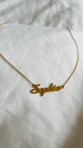 Close-up of a reviewer's gold Signature Name Necklace with the name 'Sophia' on a white fabric background.