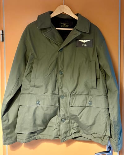 Customer's olive green US Navy AL-1 Flight Jacket on a hanger, featuring the authentic nameplate added by the reviewer.