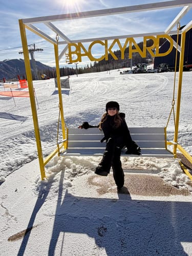 Reviewer wearing the black one-piece ski suit while sitting on a swing at a sunny ski resort.