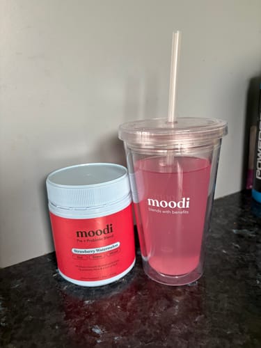 Customer's tub of Strawberry Watermelon powder next to a clear tumbler of the prepared pink drink on a counter.