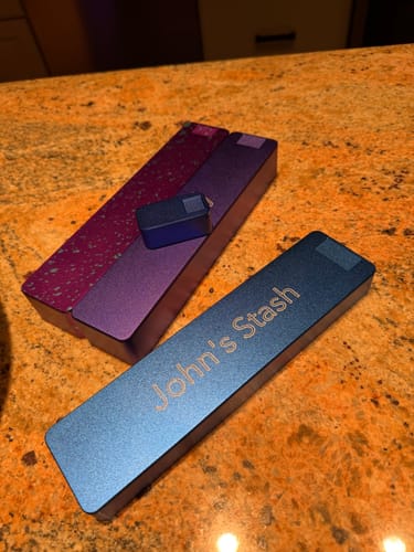 Customer's blue Weekly Case, custom engraved with 'John's Stash', shown with other cases on a counter.
