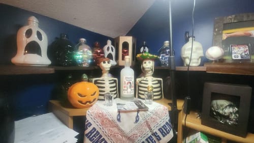 Customer's Kah Blanco Tequila bottle displayed on a shelf with a collection of skeleton figurines and other bottles.