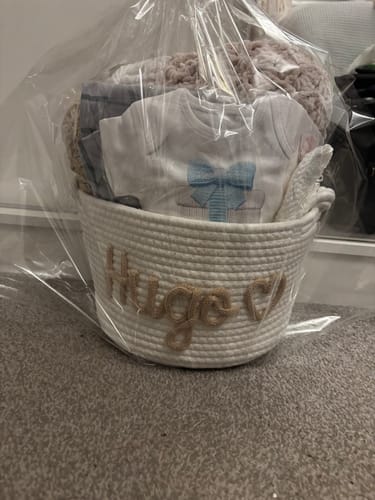 Reviewer's personalized white rope basket with the name 'Hugo', filled with baby clothes and a blanket, wrapped in cellophane.