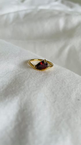 Customer's gold Pia Birthstone Ring with a pear-cut red stone, shown up close on a white fabric surface.