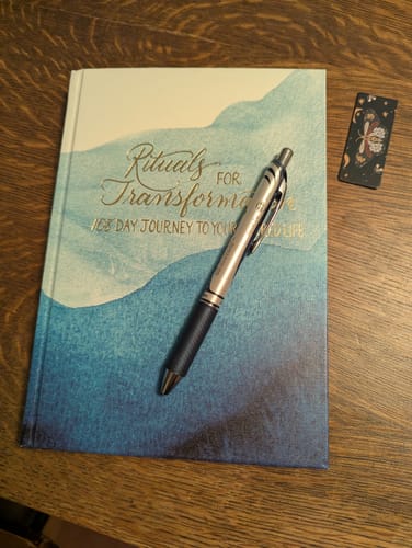 Customer's Rituals For Transformation Guided Journal with a pen on its blue watercolor cover, set on a wooden table.