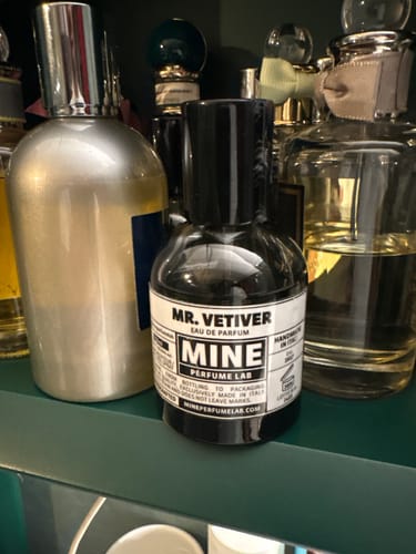 Recensore mostra il flacone di profumo MR. VETIVER su una mensola verde tra la sua collezione.