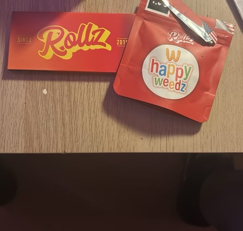 Customer shows the red pouch of Happy Weedz (CALI) THCA Flower and a matching card on a wooden table.