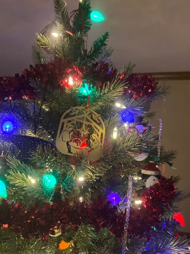 Customer's wooden Nativity Ornament hanging on a lit Christmas tree with colorful lights.