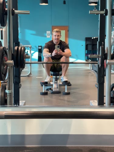 A reviewer takes a mirror selfie on a weight bench at the gym, showing their muscular arms.