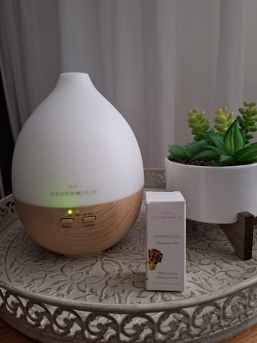 Recensent toont de Nimbus 2.0 diffuser met houtlook-basis op een dienblad, naast het doosje Happiness olie.