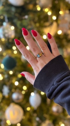 Reviewer's hand with bright red nails, wearing the Toi Et Moi Birthstone Ring with blue and yellow stones against a festive background.