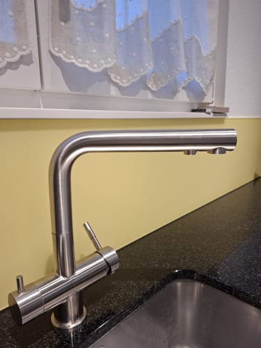 Ein Kunde zeigt die ZARA INOX 3-Wege-Armatur aus gebürstetem Edelstahl, die auf einer schwarzen Arbeitsplatte in einer Küche montiert ist.