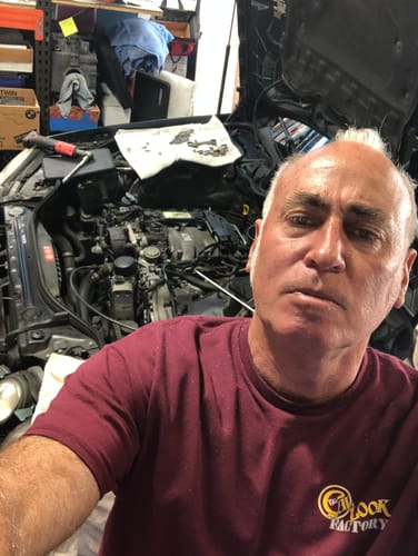 Reviewer, a mechanic, in his auto shop with a car's exposed engine in the background.