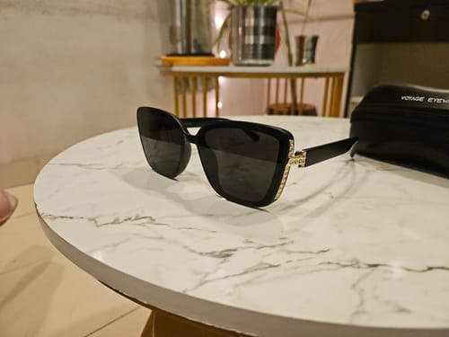 Customer's Matt Black Cateye Sunglasses with gold-tone details on the temples, resting on a marble-top table.
