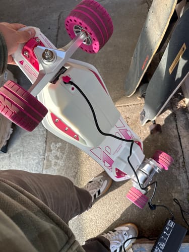Customer holding their white Meepo Voyager X electric skateboard with pink wheels as it charges on the floor.