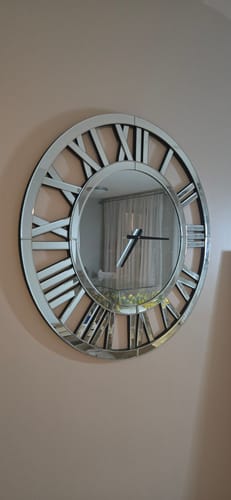 Reviewer's large Glass Mirrored Clock with Roman numerals hanging on a light-colored wall, reflecting the room.