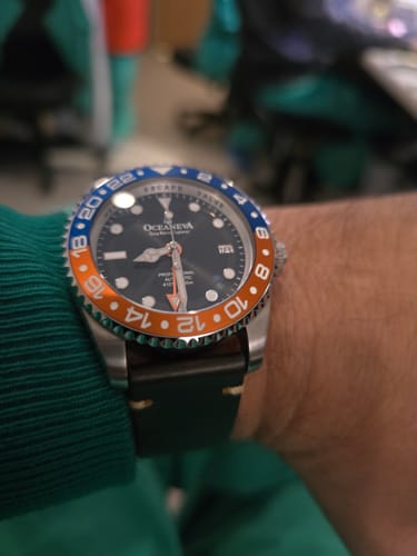 Close-up of the Oceaneva GMT watch on a reviewer's wrist, featuring its blue and orange bezel and a dark leather strap.