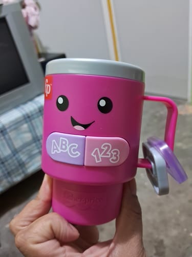 Un cliente sostiene la Taza de Café Interactiva rosa de Fisher-Price, mostrando su cara sonriente y sus botones ABC y 123.