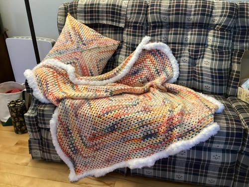 Customer's finished Hearthside Rainbow Blanket and matching pillow from the kit, displayed on a plaid armchair.