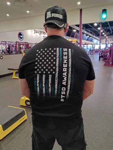 Reviewer at a gym showing the back of a black T.V.P. t-shirt with a large PTSD Awareness flag graphic.