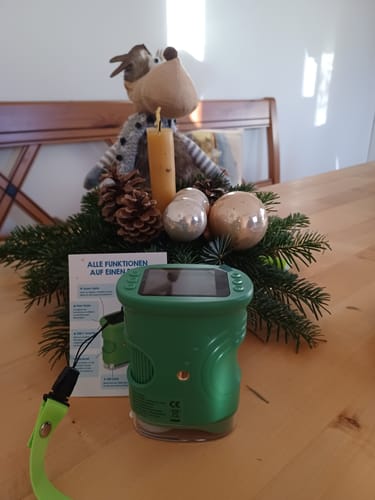 Rezensent zeigt das grüne Edubini Abenteuer-Mikroskop auf einem Tisch vor weihnachtlicher Dekoration.