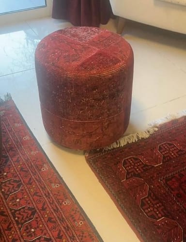 Reviewer's red handmade Hali X-Bursa Pouf sitting on a tiled floor between two traditional red rugs.