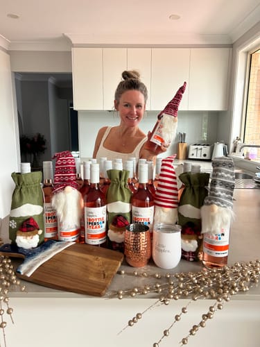 Reviewer smiling in a kitchen, holding up a bottle from the Chrissy GWP ALL wine set with a festive gnome topper.