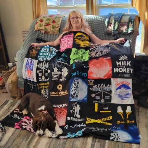 Reviewer on a couch displaying a large Premium T Shirt Quilt made of colorful dart shirts, with a dog lying at the bottom.