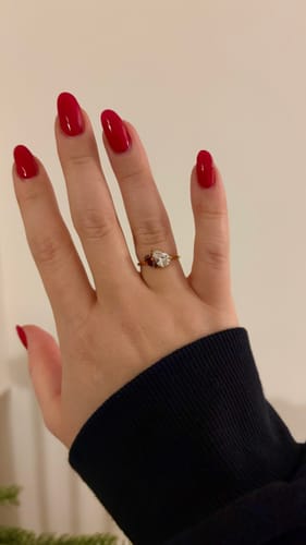 Reviewer's hand with red nails, wearing the gold Leesha Birthstone Ring with one clear and one red stone.