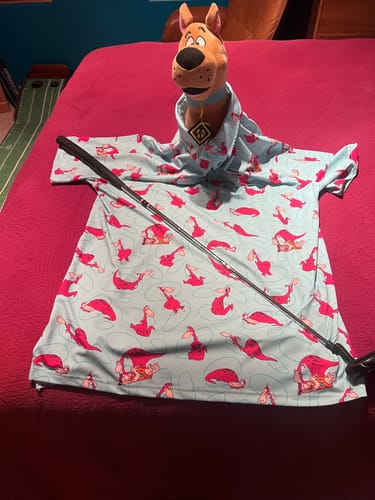 Reviewer's 'Caveman's Best Friend' polo shirt playfully modeled by a Scooby-Doo plush toy on a bed with a golf club.