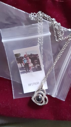 Kunde zeigt einen silberfarbenen, herzförmigen Anhänger mit Kette und ein kleines Foto in der Verpackung.
