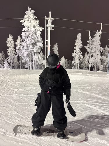 Reviewer in full black gear with the snow jacket, standing on a snowboard on a snowy hill at night.
