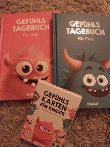 Rezensent zeigt zwei Gefühlstagebücher für Kinder in Pink und Blau, zusammen mit den Gefühlskarten auf einem Teppich.