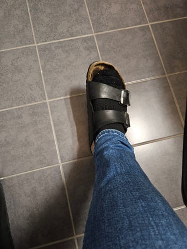 Kund som bär en svart Diasocks stödstrumpa med en sandal, sedd uppifrån på ett klinkergolv.