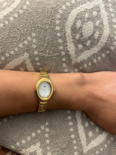 Customer wearing the elegant Classic Gold Oval Watch on their wrist against a patterned gray background.