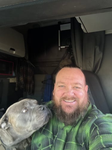 Customer smiling in a truck cab, wearing a green and black plaid flannel shirt next to his dog.