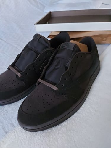 Recensent toont een paar donkerbruine Air Jordan 1 Travis Scott-sneakers zonder veters op een witte ondergrond.