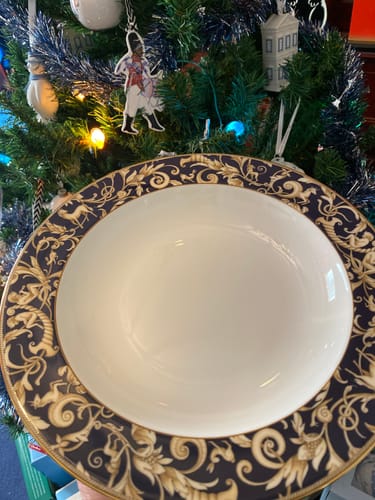 Customer holds up their Wedgwood Cornucopia Pasta Bowl, showing its wide, ornate blue and gold rim in front of a Christmas tree.