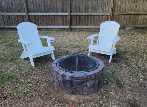 Customer's two white Folding Adirondack Chairs set up around a stone fire pit in a backyard.