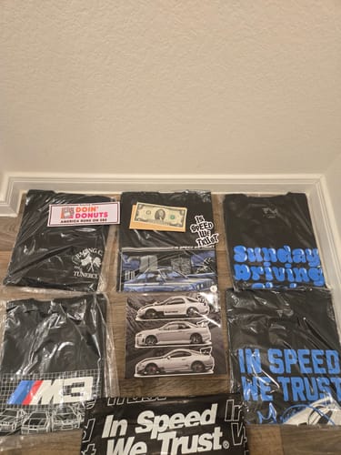 Reviewer's collection of black Mystery Tees with various car graphics, including an M3 design, laid out on a floor.