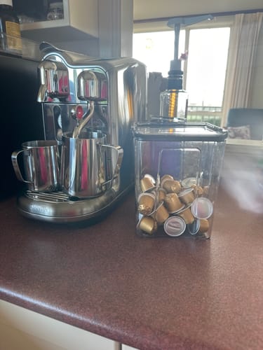 Reviewer's Blend 14 coffee pods in a clear container next to a shiny coffee machine on a countertop.