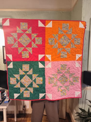 Reviewer holding up a colorful patchwork quilt made from red, orange, green, and pink polycotton fabric.